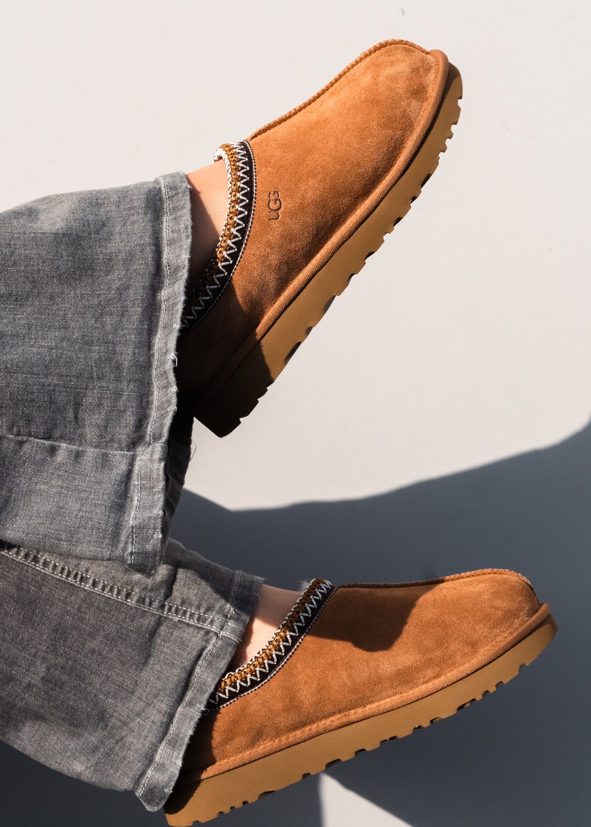 A pair of tan suede slip-on clogs with a black and white zigzag trim, shown being worn with gray jeans against a light gray background.