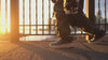 Person wearing white HOKA sneakers and dark pants while walking along a waterfront at sunset with a city skyline in the background.